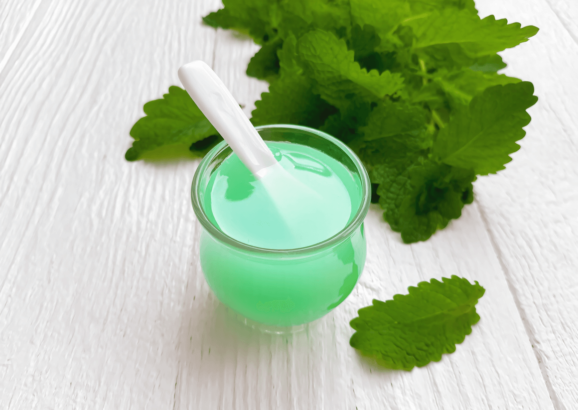 pastis&olive_sirop-de-menthe verrine de sirop vert tendre, sur table bois clair avec feuilles fraîches🔖